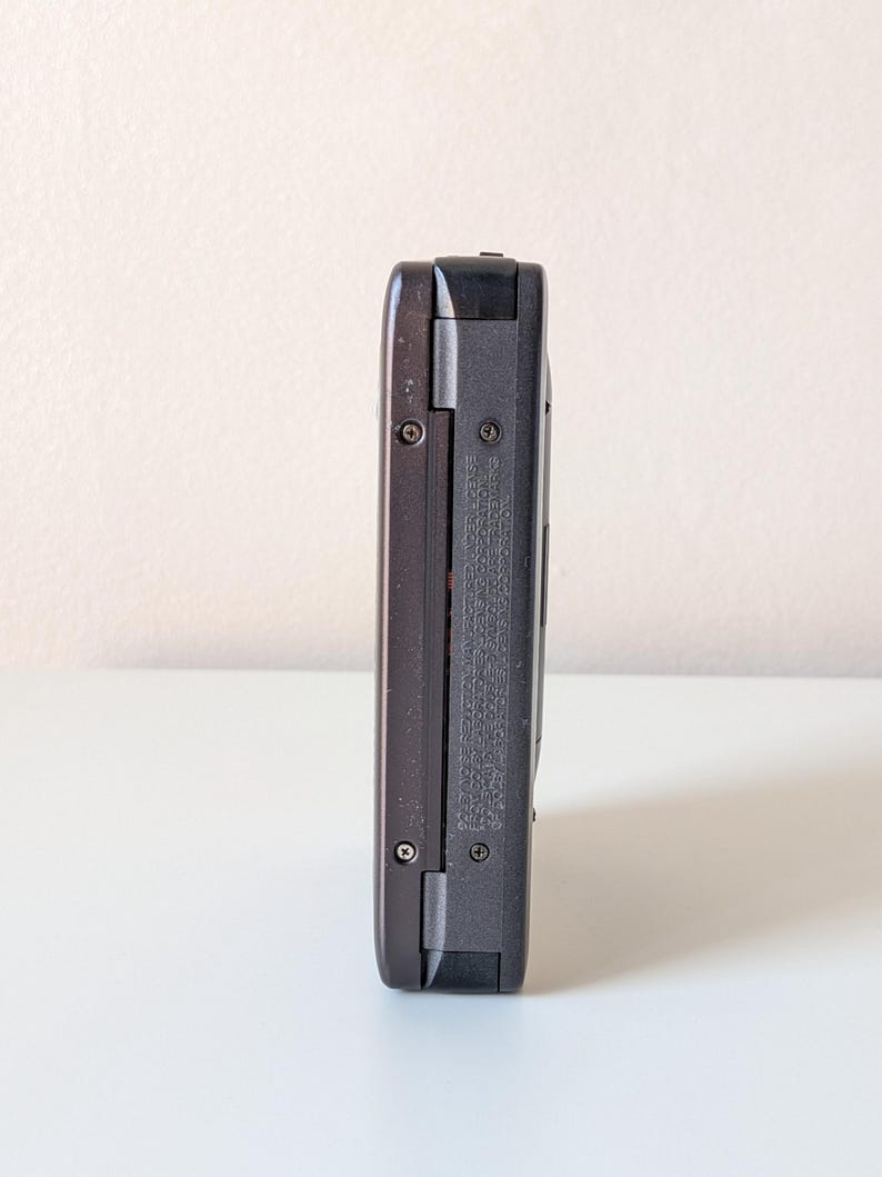 May include: A dark gray electronic device with a rectangular, elongated body. The side profile shows a textured surface with two visible metal screws and small, embossed text detailing manufacturing information and licensing specifications.