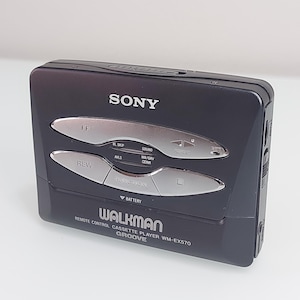 May include: A black Sony Walkman cassette player, model WM-EX570, with silver control buttons. The device has the word "WALKMAN" printed on the front, along with other text. The player is a vintage audio device.