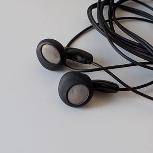 May include: A pair of black wired earbuds featuring circular housings with matte black frames and metallic silver mesh speaker grilles. The thin black audio cable is coiled behind the earbuds, connecting to a standard plug not fully visible.