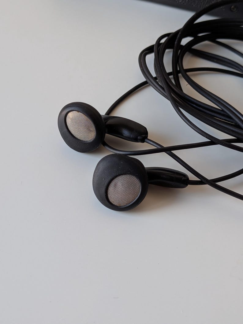 May include: A pair of black wired earbuds featuring circular housings with matte black frames and metallic silver mesh speaker grilles. The thin black audio cable is coiled behind the earbuds, connecting to a standard plug not fully visible.