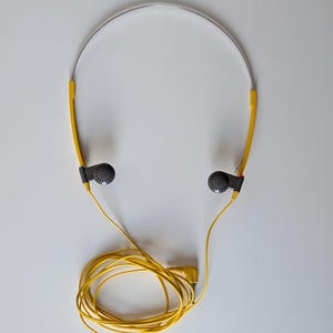 May include: Lightweight headphones featuring a thin silver metal headband, bright yellow plastic side arms, and small dark gray circular earpieces. A long yellow cord is attached to the earpieces, ending in a standard yellow audio jack plug.