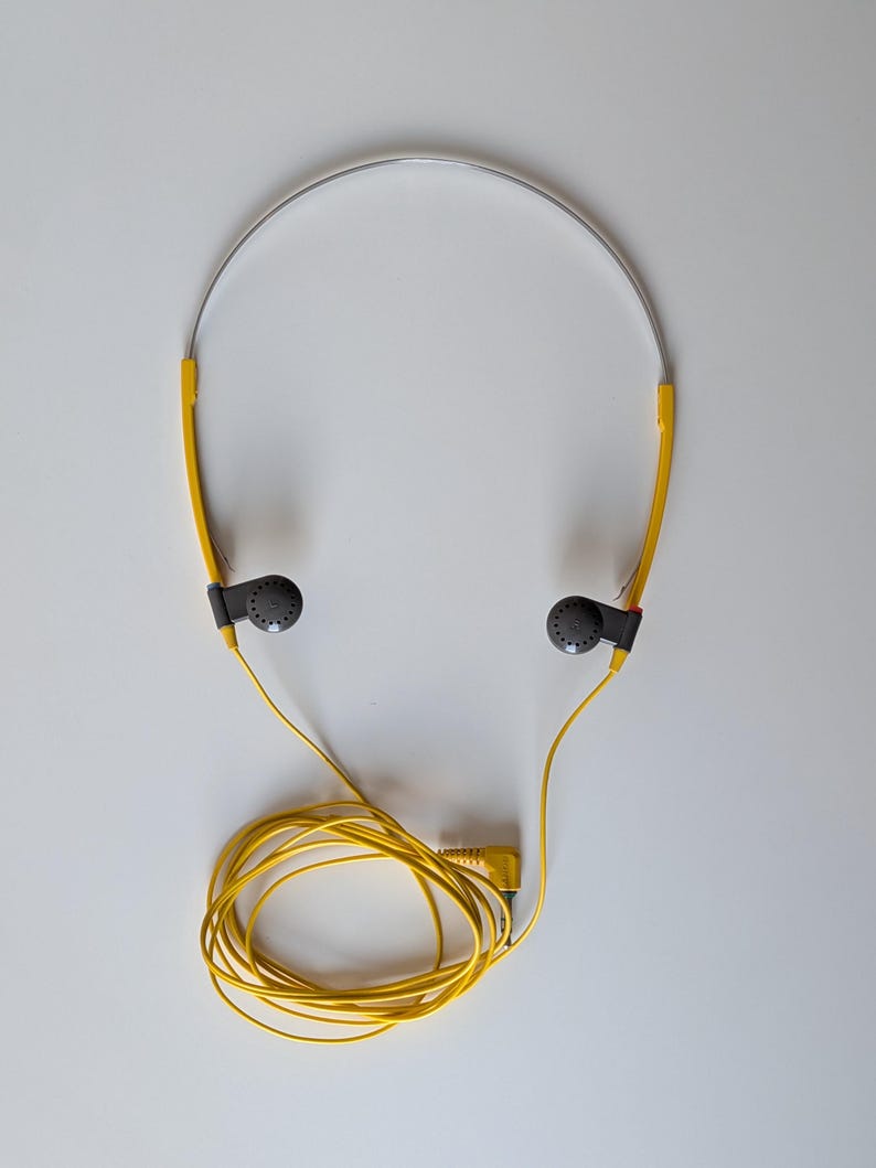 May include: Lightweight headphones featuring a thin silver metal headband, bright yellow plastic side arms, and small dark gray circular earpieces. A long yellow cord is attached to the earpieces, ending in a standard yellow audio jack plug.