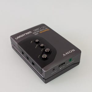 May include: A grey Sony Walkman cassette player with black control buttons. The device has the word "SONY" printed on the side and "WALKMAN" on the top. It features a headphone socket and volume control.