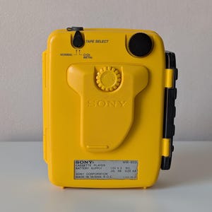 May include: A bright yellow Sony Walkman cassette player, model WM-B53, featuring a central belt clip, a black volume dial, and a tape select switch for Normal, CrO2, and Metal tapes. The device includes a label with technical specifications.