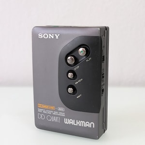 May include: A grey Sony DD Quartz Walkman portable cassette player. The device has a black control panel with play, stop, rewind, and fast forward buttons. The text "SONY" is printed at the top, and "DD QUARTZ WALKMAN" is printed at the bottom.