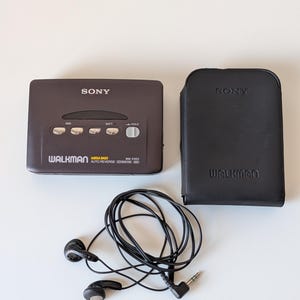 May include: A dark gray Sony Walkman WM-EX53 cassette player with silver control buttons, a black leather carrying case embossed with the Sony and Walkman logos, and a pair of black wired earbuds with a standard 3.5mm audio jack connector.