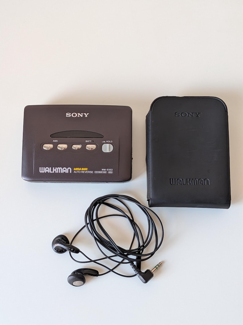 May include: A dark gray Sony Walkman WM-EX53 cassette player with silver control buttons, a black leather carrying case embossed with the Sony and Walkman logos, and a pair of black wired earbuds with a standard 3.5mm audio jack connector.