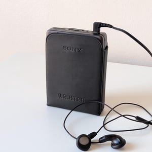 May include: A black Sony Walkman portable cassette player encased in a black leather-like protective cover. The device features the embossed text SONY and WALKMAN on the front and is connected to a pair of black wired earbuds via a thin black cord.