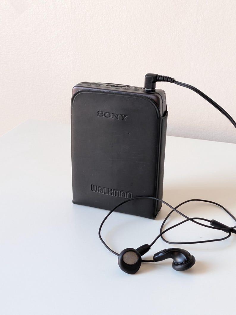 May include: A black Sony Walkman portable cassette player encased in a black leather-like protective cover. The device features the embossed text SONY and WALKMAN on the front and is connected to a pair of black wired earbuds via a thin black cord.