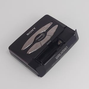 May include: Black Sony Walkman WM-EX570 cassette player with silver control buttons. The portable audio device has a battery compartment and the Walkman logo is visible. The device is on a white background.