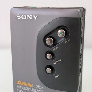 May include: A grey Sony Walkman cassette player with a black control panel. The player has buttons for play, stop, rewind, and fast forward. The text "DD QUARTZ WALKMAN" is visible. It also features "DOLBY B NR" and "AVLS" logos.