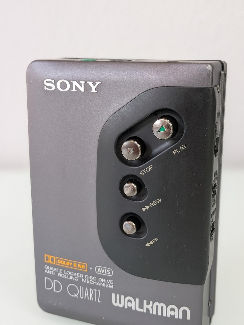 May include: A grey Sony Walkman cassette player with a black control panel. The player has buttons for play, stop, rewind, and fast forward. The text "DD QUARTZ WALKMAN" is visible. It also features "DOLBY B NR" and "AVLS" logos.