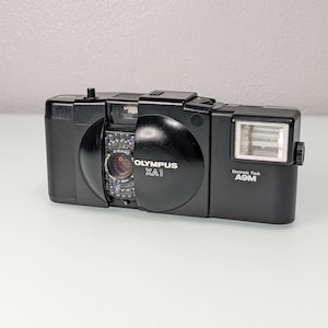 Può includere: Fotocamera Olympus XA1 nera con flash integrato. La fotocamera presenta la scritta "Olympus XA1" sul davanti e "Electronic Flash A9M" sul lato. L'obiettivo è visibile e riporta la scritta "D.Zuiko".