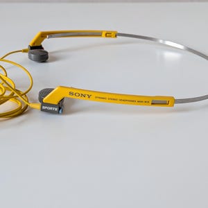 May include: Yellow and grey Sony Dynamic Stereo Headphones MDR-W15 with a thin metal headband. The yellow plastic frame features the text SONY, DYNAMIC STEREO HEADPHONES MDR-W15, and SPORTS. A yellow cord is attached to the grey ear pieces.
