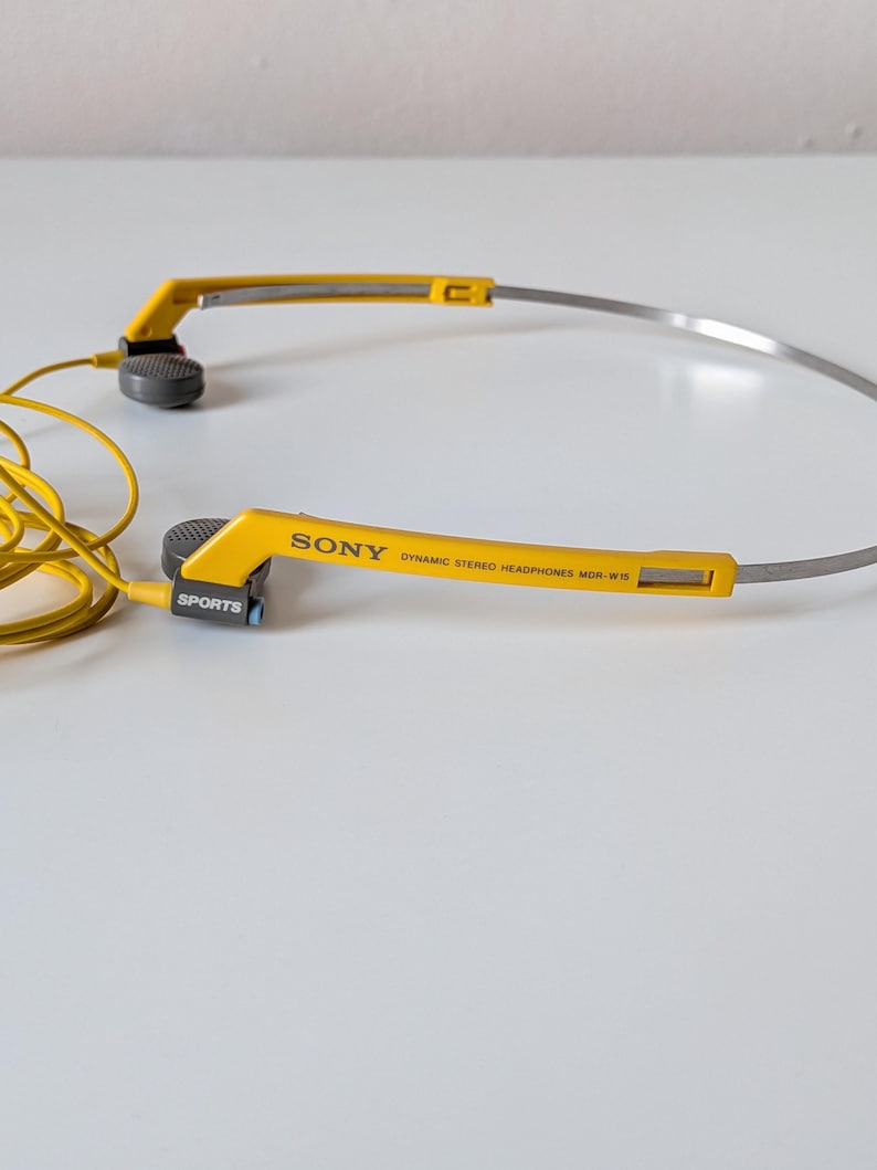 May include: Yellow and grey Sony Dynamic Stereo Headphones MDR-W15 with a thin metal headband. The yellow plastic frame features the text SONY, DYNAMIC STEREO HEADPHONES MDR-W15, and SPORTS. A yellow cord is attached to the grey ear pieces.