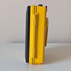 May include: A rectangular protective case featuring a bright yellow exterior shell and a contrasting black lid. The side profile displays a sturdy hinge mechanism, a secure latching system, and a molded handle attached to the yellow body section.