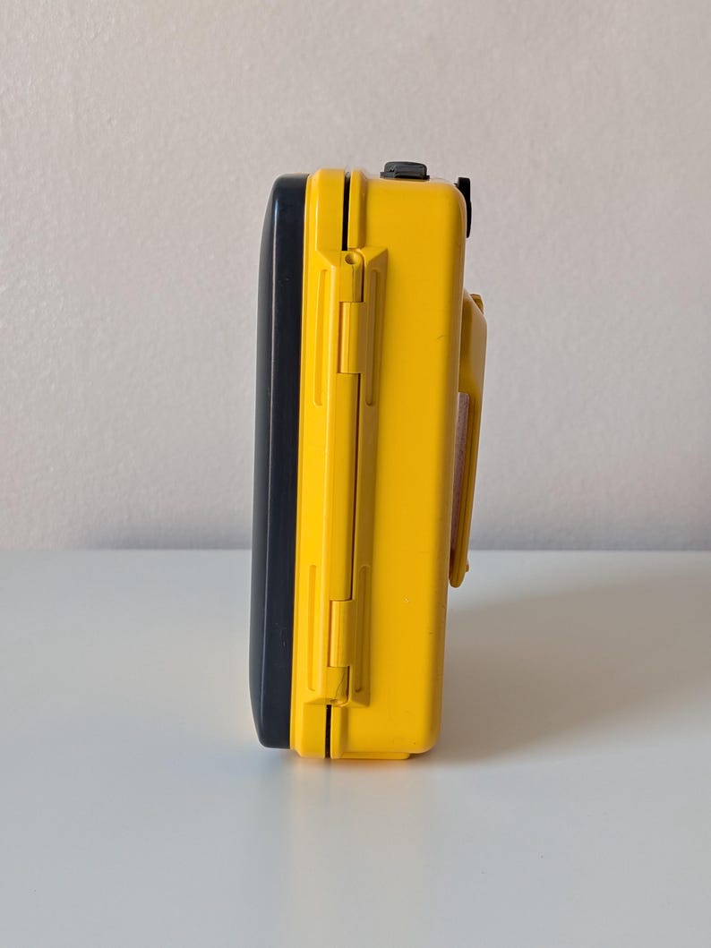 May include: A rectangular protective case featuring a bright yellow exterior shell and a contrasting black lid. The side profile displays a sturdy hinge mechanism, a secure latching system, and a molded handle attached to the yellow body section.