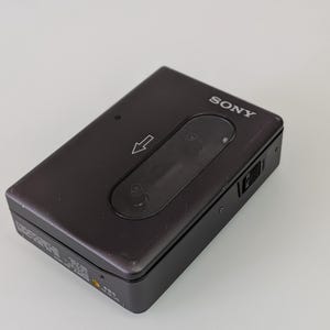 May include: A dark grey Sony Walkman cassette player. The player has a rectangular shape with rounded corners and a clear window. The Sony logo is visible on the top right corner. A directional arrow is printed on the front.