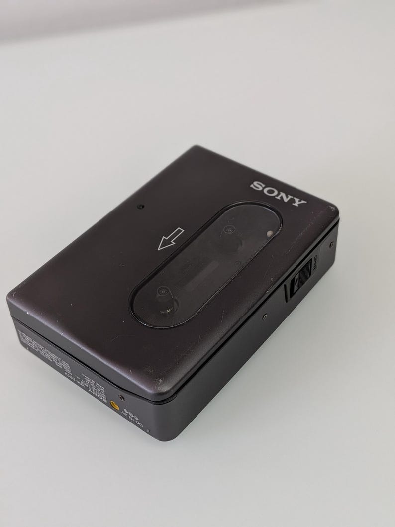 May include: A dark grey Sony Walkman cassette player. The player has a rectangular shape with rounded corners and a clear window. The Sony logo is visible on the top right corner. A directional arrow is printed on the front.