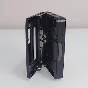 May include: Black portable cassette player with the cassette door open. The player is on a white surface. The cassette compartment is open, revealing the internal components. The player has a silver and black design.