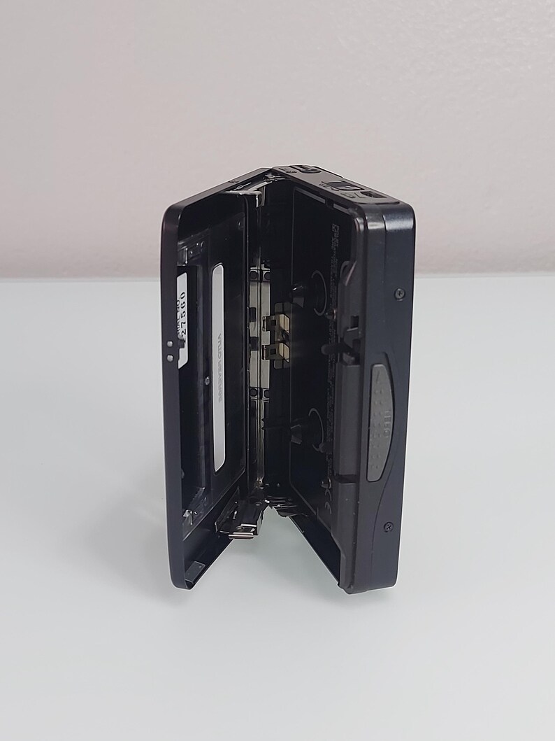 May include: Black portable cassette player with the cassette door open. The player is on a white surface. The cassette compartment is open, revealing the internal components. The player has a silver and black design.