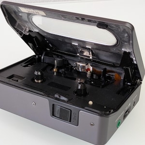 May include: A grey portable cassette player with the top open, revealing the internal components. The player has a clear plastic window and a black interior. The "OPEN" button is visible on the front. The device has a headphone socket and a battery indicator.