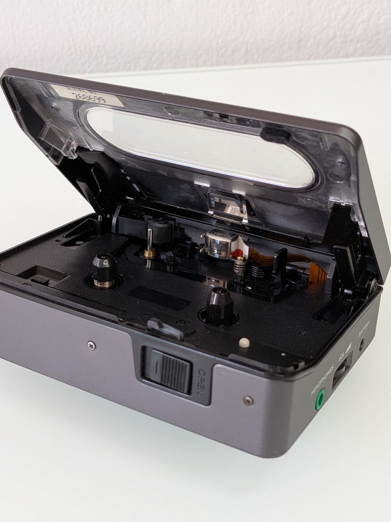 May include: A grey portable cassette player with the top open, revealing the internal components. The player has a clear plastic window and a black interior. The "OPEN" button is visible on the front. The device has a headphone socket and a battery indicator.