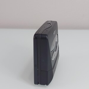 May include: A black Sony Walkman cassette player, seen from the side. The device has several buttons and a small wheel on the top. The Sony logo is visible on the front. The player is rectangular with rounded edges.
