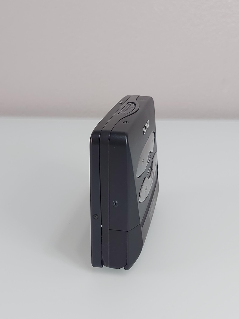 May include: A black Sony Walkman cassette player, seen from the side. The device has several buttons and a small wheel on the top. The Sony logo is visible on the front. The player is rectangular with rounded edges.