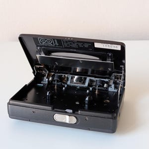 May include: A dark gray portable cassette player with its lid open, revealing the internal tape mechanism, two spindles, and a silver latch. A label inside reads: Note: To enjoy optimum stereo sound, clean the head, the capstans and the pinch rollers.