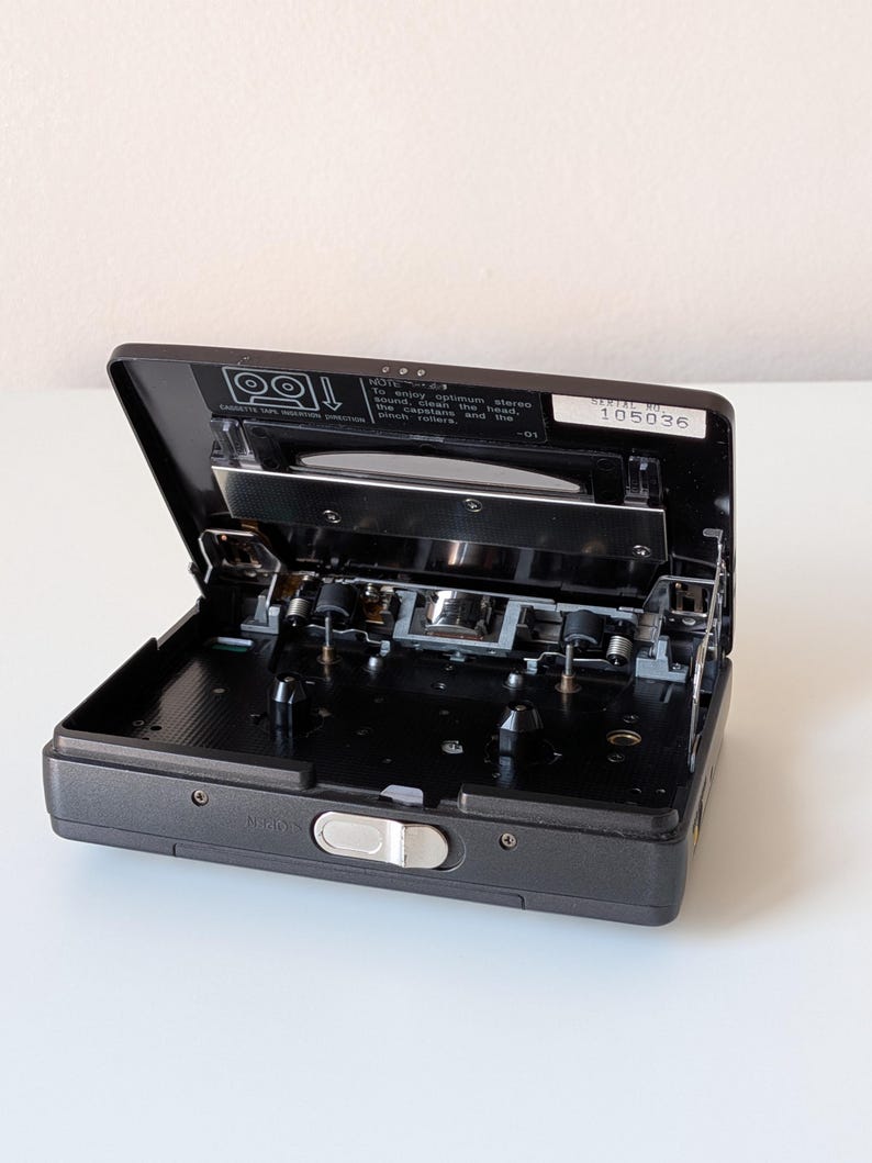 May include: A dark gray portable cassette player with its lid open, revealing the internal tape mechanism, two spindles, and a silver latch. A label inside reads: Note: To enjoy optimum stereo sound, clean the head, the capstans and the pinch rollers.