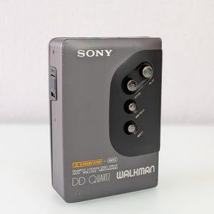 May include: A grey Sony Walkman DD Quartz portable cassette player. The device has a black control panel with play, stop, and rewind buttons. The text "SONY" is above the control panel. The Walkman has "Dolby B NR" and "AVLS" features.