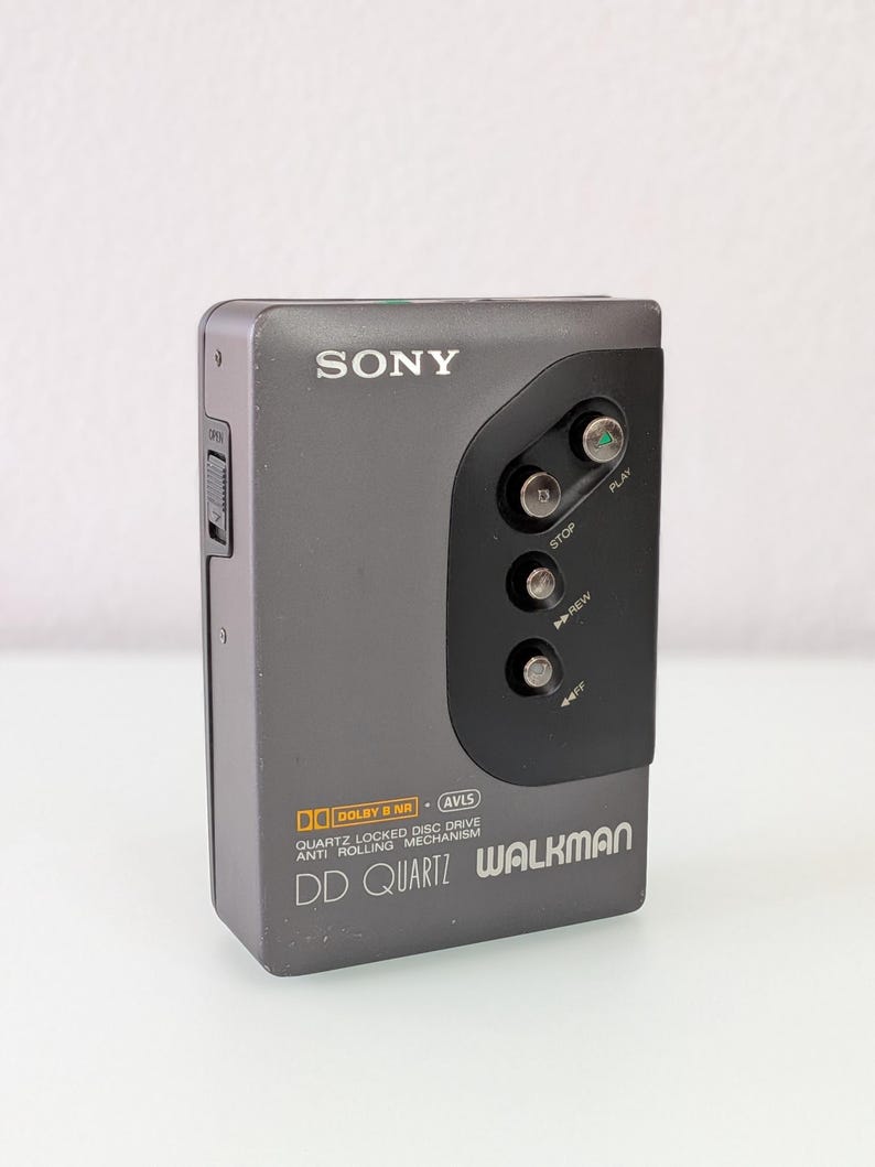 May include: A grey Sony Walkman DD Quartz portable cassette player. The device has a black control panel with play, stop, and rewind buttons. The text "SONY" is above the control panel. The Walkman has "Dolby B NR" and "AVLS" features.