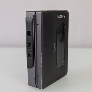 May include: A black Sony Walkman cassette player. The device has a rectangular shape with a clear window and a control panel on the side. The Sony logo is visible on the front. The arrow indicates the direction of the cassette.