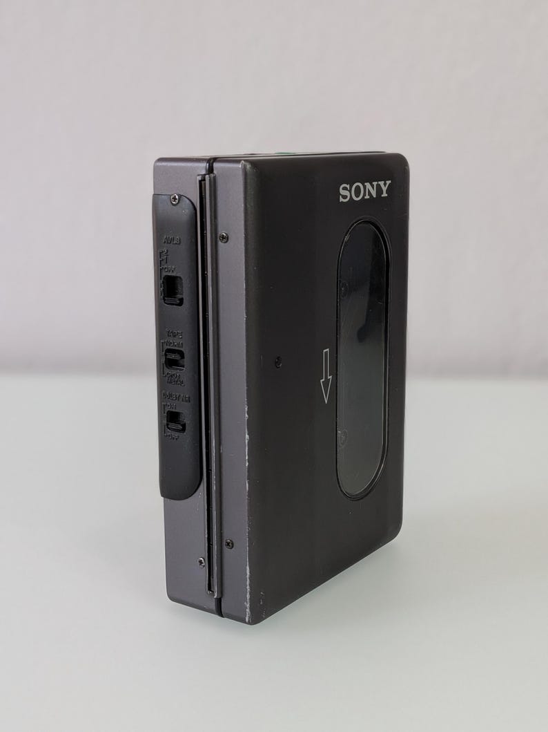 May include: A black Sony Walkman cassette player. The device has a rectangular shape with a clear window and a control panel on the side. The Sony logo is visible on the front. The arrow indicates the direction of the cassette.