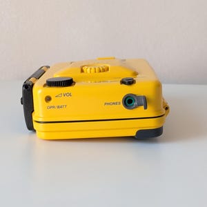 May include: A bright yellow portable cassette player featuring a black volume dial, a headphone jack labeled PHONES, and an OPR/BATT indicator light. The device has a rectangular shape with black accents and clearly printed text labels for controls.