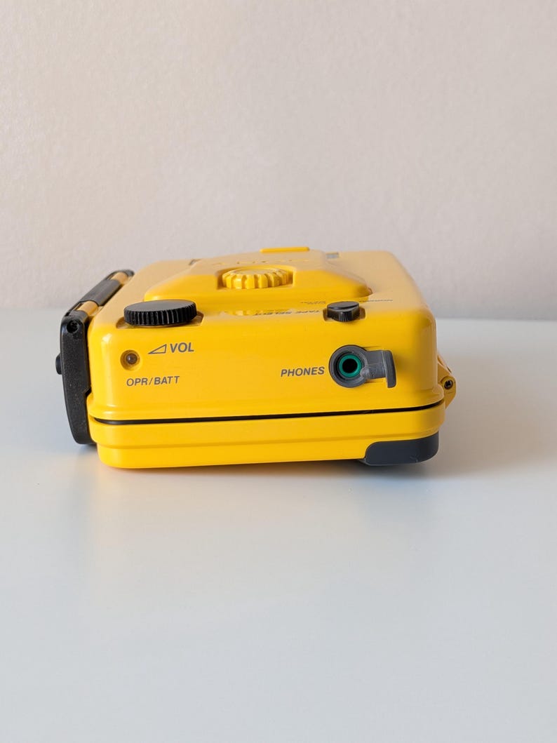 May include: A bright yellow portable cassette player featuring a black volume dial, a headphone jack labeled PHONES, and an OPR/BATT indicator light. The device has a rectangular shape with black accents and clearly printed text labels for controls.