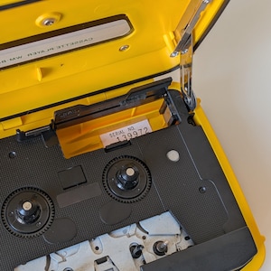 May include: A bright yellow portable cassette player with its lid open, revealing the internal black mechanism, two circular spindles, and a white label with the text SERIAL NO. 139972. The interior lid features the text CASSETTE PLAYER.