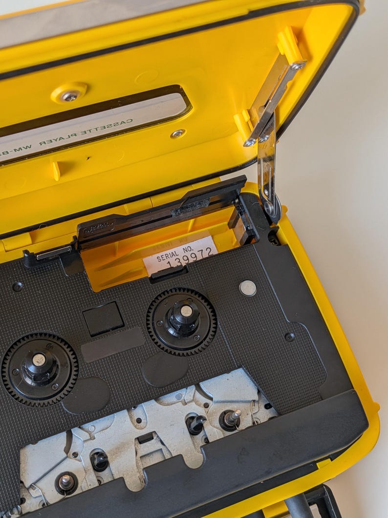 May include: A bright yellow portable cassette player with its lid open, revealing the internal black mechanism, two circular spindles, and a white label with the text SERIAL NO. 139972. The interior lid features the text CASSETTE PLAYER.