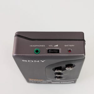 May include: A dark grey Sony Walkman cassette player. It features a headphone jack, volume control, and a battery indicator. The word "SONY" is printed on the front. There are also buttons for play, stop, and rewind.