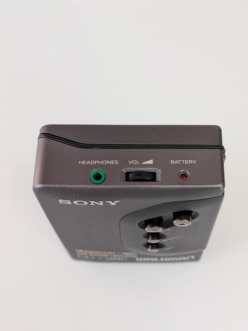 May include: A dark grey Sony Walkman cassette player. It features a headphone jack, volume control, and a battery indicator. The word "SONY" is printed on the front. There are also buttons for play, stop, and rewind.