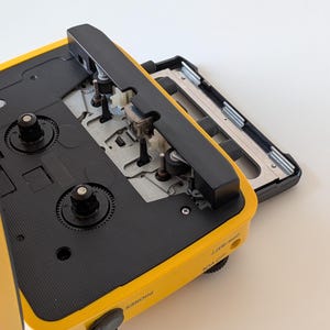 May include: A yellow portable cassette player with the cassette compartment door open, revealing the internal mechanical components, including the tape heads and spindles. The side of the device features labels for PHONES, VOL, and OPR/BATT.