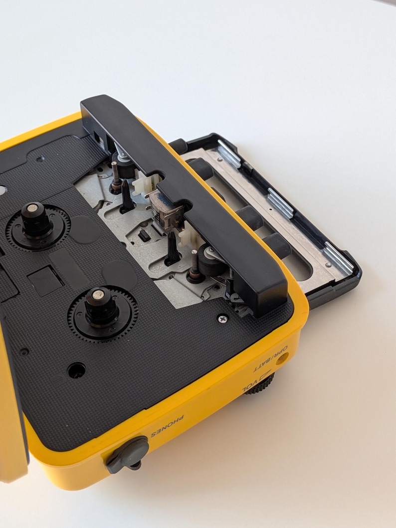 May include: A yellow portable cassette player with the cassette compartment door open, revealing the internal mechanical components, including the tape heads and spindles. The side of the device features labels for PHONES, VOL, and OPR/BATT.