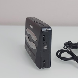 May include: A black Sony Walkman cassette player with silver buttons and the brand name visible. The device has a headphone jack and a volume control slider on the side. A black cord is visible in the background.