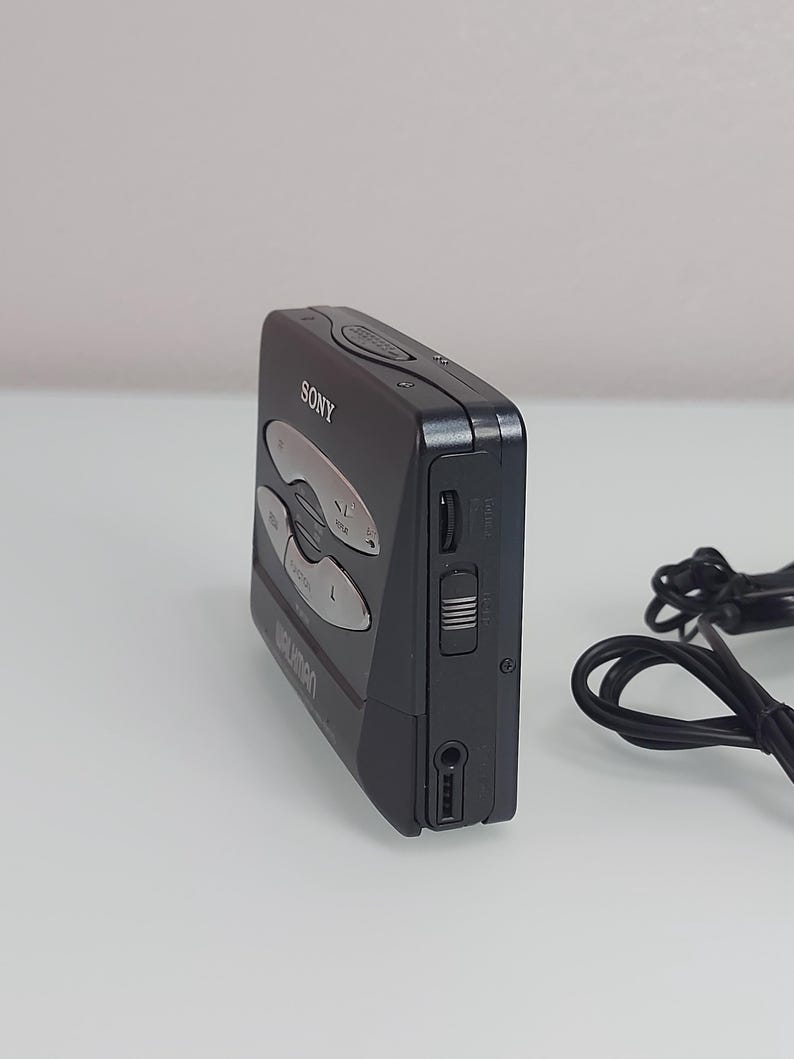 May include: A black Sony Walkman cassette player with silver buttons and the brand name visible. The device has a headphone jack and a volume control slider on the side. A black cord is visible in the background.