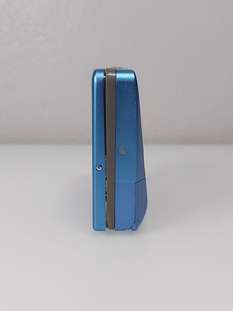 以下が含まれることがあります： 長方形の青い電子機器。デバイスはメタリック仕上げで、中央に濃い灰色のストライプがあります。デバイスは白い表面に直立しています。