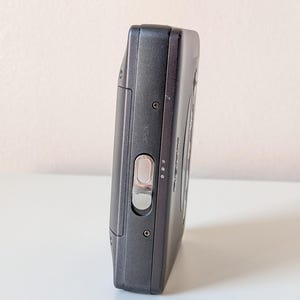 May include: A dark gray portable cassette player featuring a metallic silver sliding latch labeled OPEN on the side panel. The device has a rectangular body with visible screws and a smooth matte finish, showcasing a compact design for audio playback.