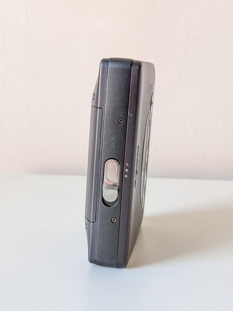 May include: A dark gray portable cassette player featuring a metallic silver sliding latch labeled OPEN on the side panel. The device has a rectangular body with visible screws and a smooth matte finish, showcasing a compact design for audio playback.