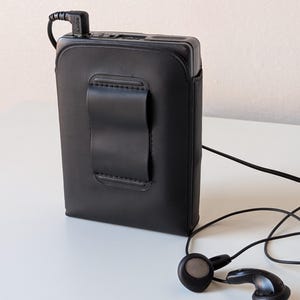 May include: A black rectangular portable cassette player encased in a fitted black leather-like protective cover with a belt clip on the back. A black Sony-branded headphone jack is plugged into the top, connected to black wired earbuds.
