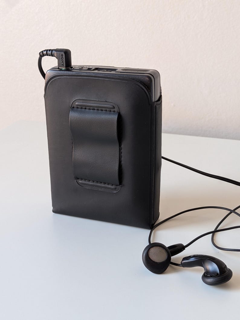 May include: A black rectangular portable cassette player encased in a fitted black leather-like protective cover with a belt clip on the back. A black Sony-branded headphone jack is plugged into the top, connected to black wired earbuds.